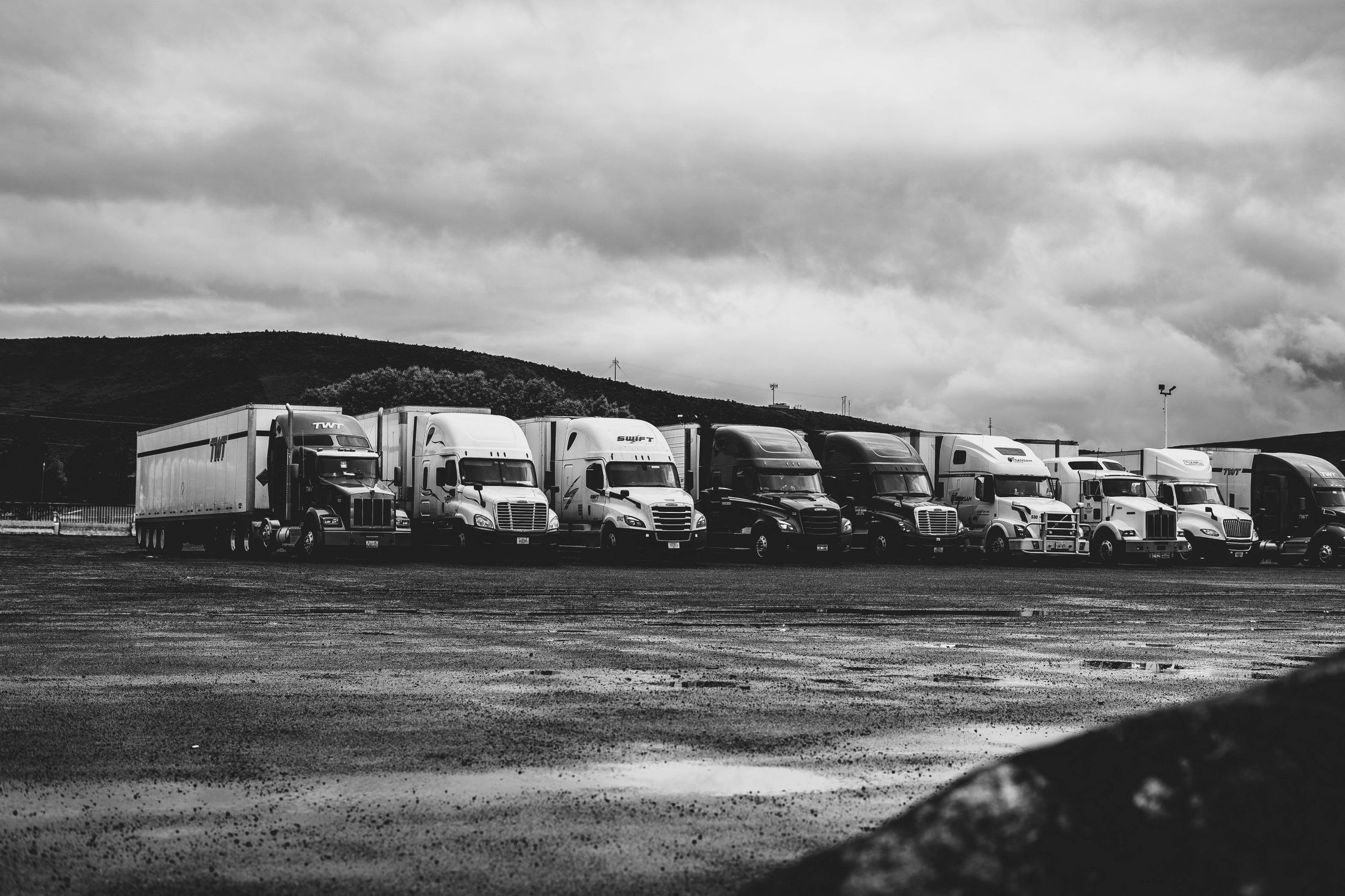 A row of conventional American semi-trucks parked in a wet lot, photographed in black and white