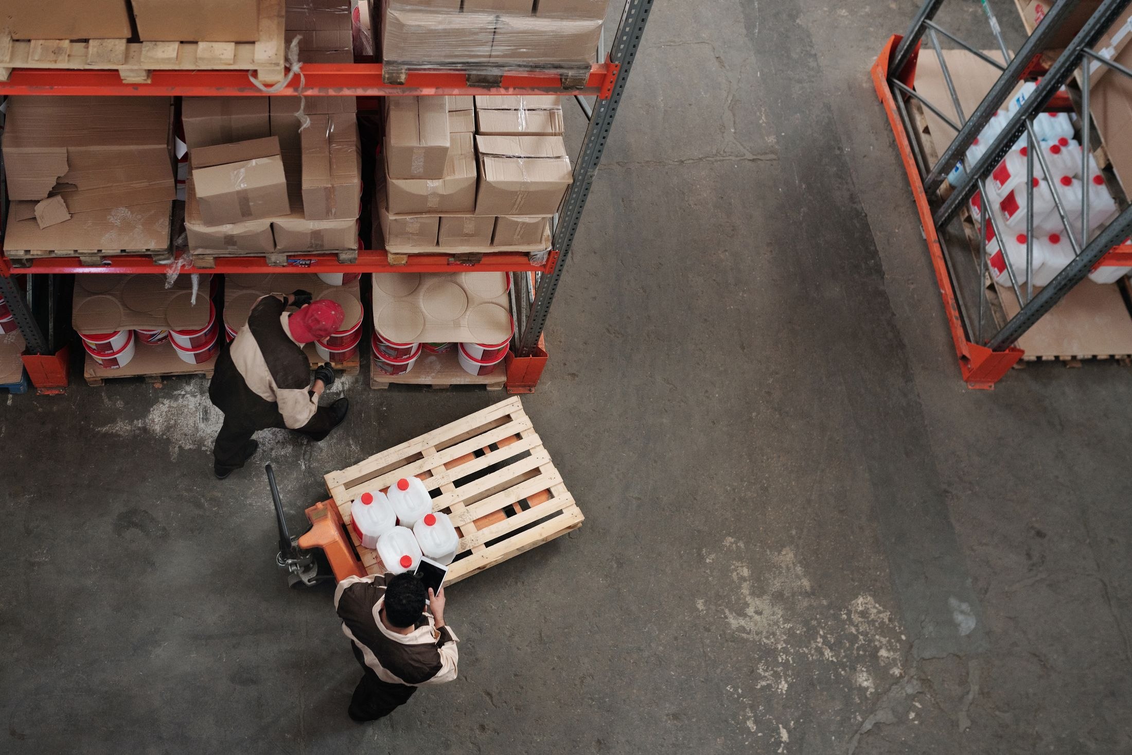 Overhead view of two warehouse workers beside storage racks and a loaded pallet jack
